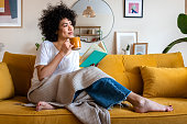 Pensive relaxed African american woman reading a book at home, drinking coffee sitting on the couch. Copy space.