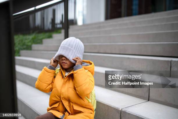 happy multiracial boy sitting on stairs with cap on his eyes in winter in town. - sad kid in kindergarten stock pictures, royalty-free photos & images