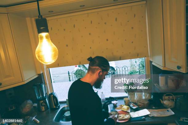 a bearded man with long curly hair in a bun is busy cooking in the kitchen, standing by the window. breakfast at home on the day off - summer hair bun stock pictures, royalty-free photos & images