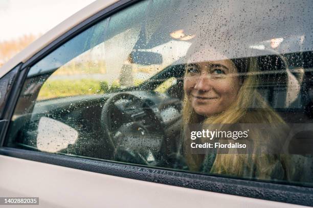 laughing woman driving car smiling through closed vehicle window. rainy day, dew and water droplets on glass, condensation - winter car window foto e immagini stock