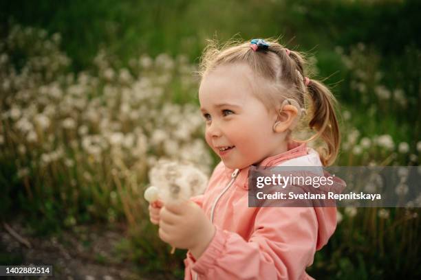 smiling toddler girl with cochlear implants walking in the park with a bouquet of dandelions - cochlea implant stock pictures, royalty-free photos & images
