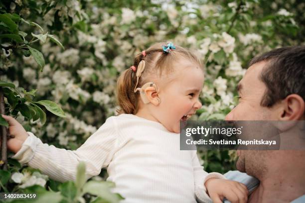 dad holds in his arms smiling joyful baby daughter with cochlear implants, in a blooming apple tree, aroma of fresh flowers, outdoor nature - cochlea stock pictures, royalty-free photos & images