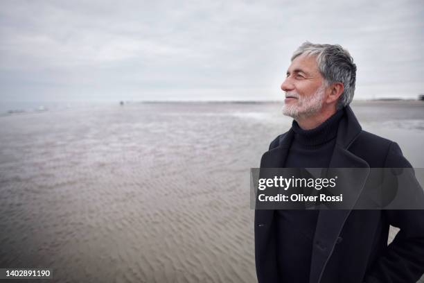 mature man in coat on the beach looking at view - man coat stock pictures, royalty-free photos & images