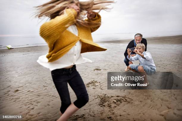 boy taking smartphone picture of girl turning around on the beach - teen boy barefoot stock-fotos und bilder
