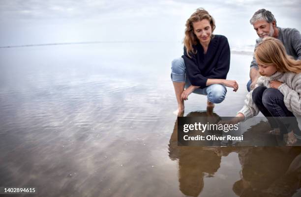 man and woman watching girl crouching in the sea touching water surface - mother and child in water at beach stock pictures, royalty-free photos & images