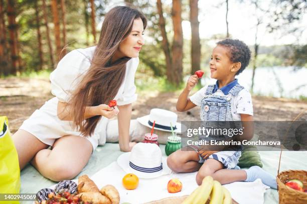 a 4-year-old boy in nature by the river with his mother on a picnic eating berries and fruits - 5 year stock pictures, royalty-free photos & images
