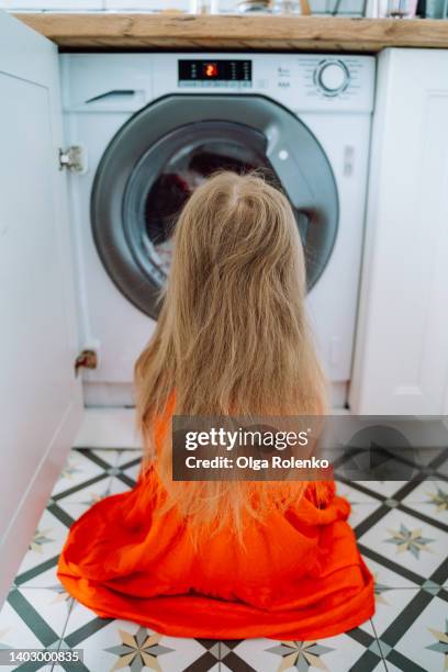 small little blond girl in red dress watching at drum of washing machine like tv. rearview - blond hair girl fotografías e imágenes de stock