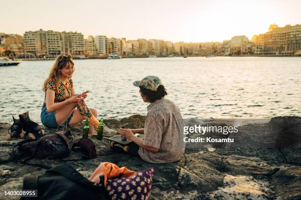 two cheerful skaters sitting on a rocky shore, smoking a joint and drinking a cold, refreshing beer - competition law stock pictures, royalty-free photos & images