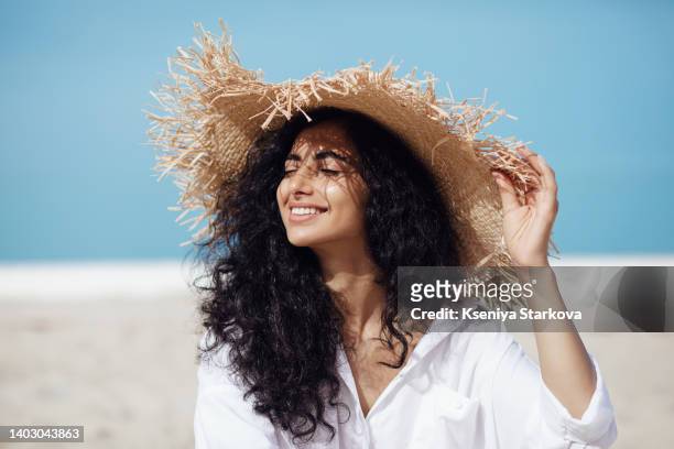 young beautiful mixed race woman with long curly hair sits on the beach in a straw hat with her eyes closed, looks at the sun and smiles - beautiful armenian women stock pictures, royalty-free photos & images