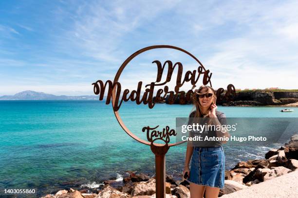 portrait of a woman at the sign that indicates the mediterranean sea at the isla de las palomas in tarifa, cadiz. - tarifa moors stock pictures, royalty-free photos & images