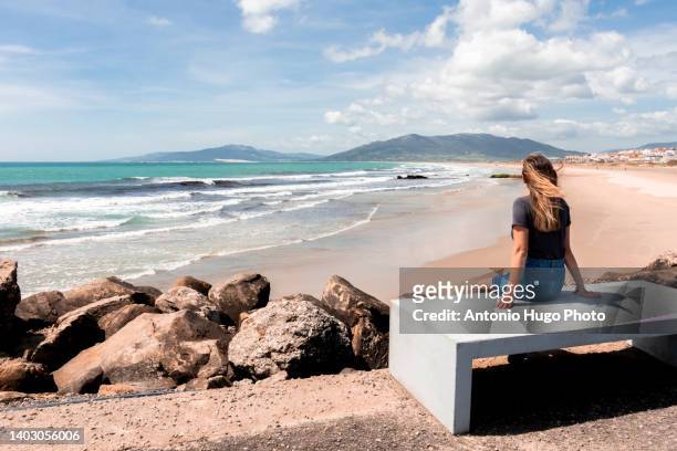 blonde woman with long hair sitting on a bench and looking at a beach. - tarifa moors stock pictures, royalty-free photos & images