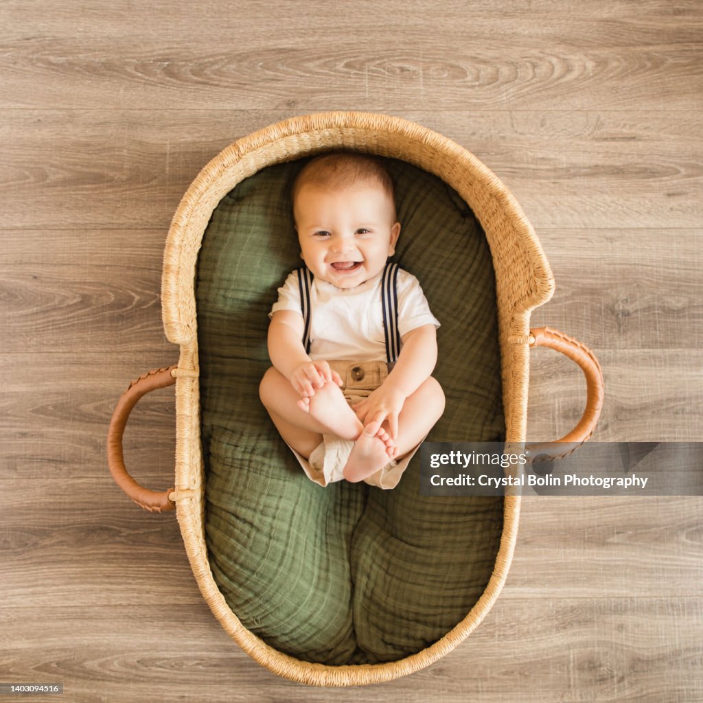A 25-Week-Old Baby Boy Holding His Feet, Wearing a White Shirt With Navy Striped Suspenders & Khaki Shorts While Laying in a Cozy Green Cotton Blanket in a Seagrass Moses Basket Surrounded by Tropical House Plants for a Boho Tropical Vibe