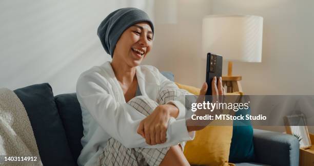 junge asiatische frau, die an krebs erkrankt ist, telefoniert mit freunden, die zu hause auf dem sofa im wohnzimmer sitzen. - cellphone cancer illness stock-fotos und bilder