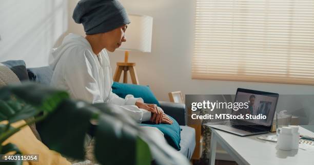 young asian woman sick with cancer using laptop talk to doctor sitting on sofa in living room at home. - mental health services stock pictures, royalty-free photos & images