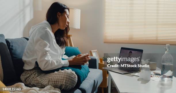 sick young asian woman using laptop talk to doctor sitting on sofa in living room at home. - telemedicine stock pictures, royalty-free photos & images
