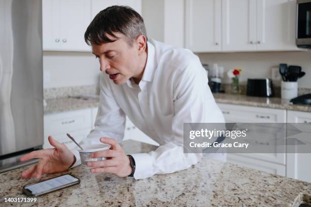 man snacks on cup of yogurt while using smart phone in kitchen - joghurtbecher stock-fotos und bilder