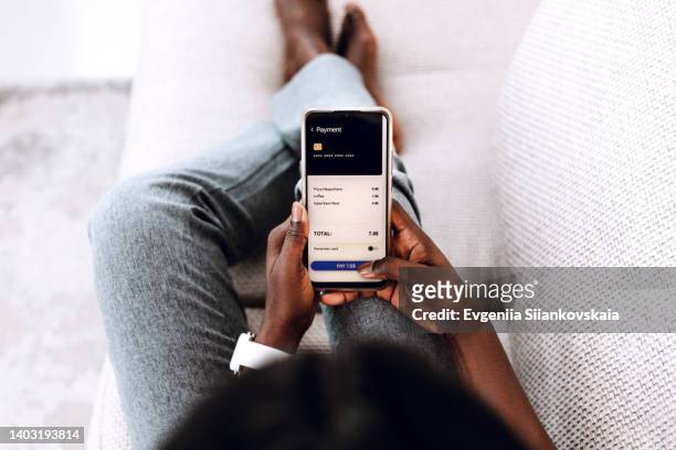 over the shoulder view of young black woman making order in cafe with smartphone sitting on the sofa at home. - frau mit handy screen stock-fotos und bilder