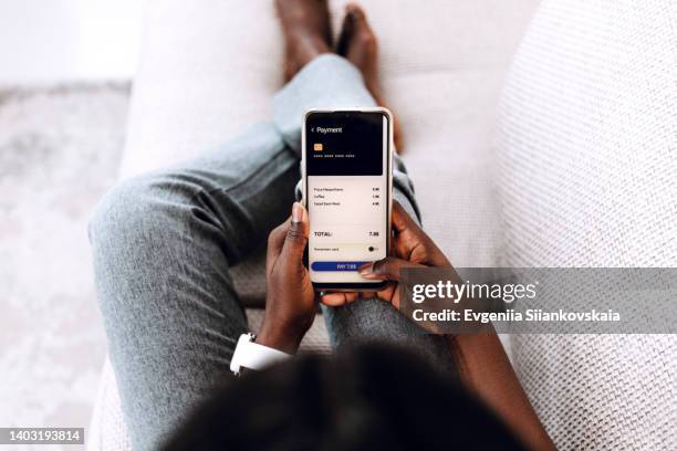 over the shoulder view of young black woman making order in cafe with smartphone sitting on the sofa at home. - pago por móvil fotografías e imágenes de stock