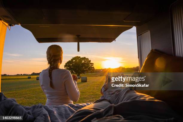 man lying down in his van with outstretched legs, back door of van is open, women with blond hair standing outside, holding a flower, view of an agricultural field with hay bales during sunset - woman stretching sunset stockfoto's en -beelden