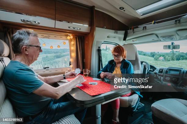 old senior couple, man with gray hair and women with dyed red hair, both wearing eyeglasses, sitting inside their camper van and playing cards - senior colored hair stock pictures, royalty-free photos & images