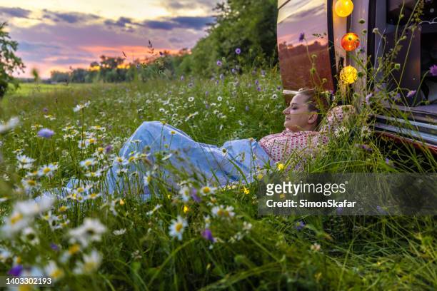 unbeschwerte frau, die sich über gras inmitten von pflanzen vor dem wohnmobilanhänger gegen den himmel entspannt - frau blumenwiese stock-fotos und bilder