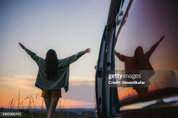 woman with brown hair with brown hair and outstretched arms walking next to her van in the nature during sunset - car sunset arm stock pictures, royalty-free photos & images