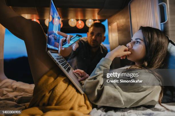 woman with brown hair with brown hair lying down in her van, working on her laptop, her boyfriend standing outdoors, holds a smartphone and pointing on something on the screen - camping indoors stock pictures, royalty-free photos & images