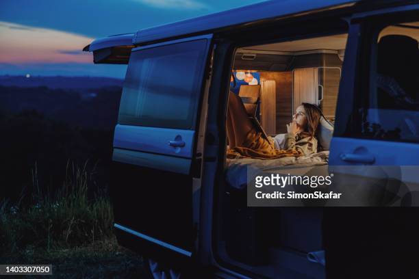 woman with brown hair lying down in her van, working on her laptop in the evening, legs outstretched, doors of van opened, view from outdoors - vehicle door stock pictures, royalty-free photos & images