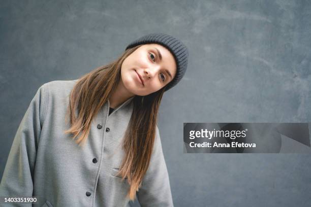 beautiful smiling woman with gray eyes in gray coat and gray knitted cap stands against gray wall outdoors. backdrop for your design with copy space. front view - gray hat stockfoto's en -beelden