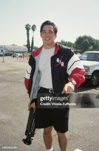 American actor Dean Cain, wearing a black, red and white Air Jordan jacket over a grey t-shirt with black shorts, carrying a tripod in his right...