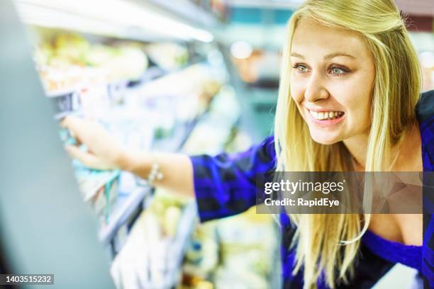 young blonde woman shopping in supermarket reaches for a dairy product on refrigerated shelves, smiling happily - mejeriavdelning bildbanksfoton och bilder