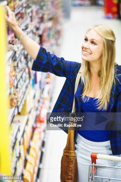 glückliche junge blonde frau, die im supermarkt einkauft, reicht bis zu einem hohen regal, wenn sie sich für einen kauf entscheidet - erwachsene im geschäft in höherer position stock-fotos und bilder