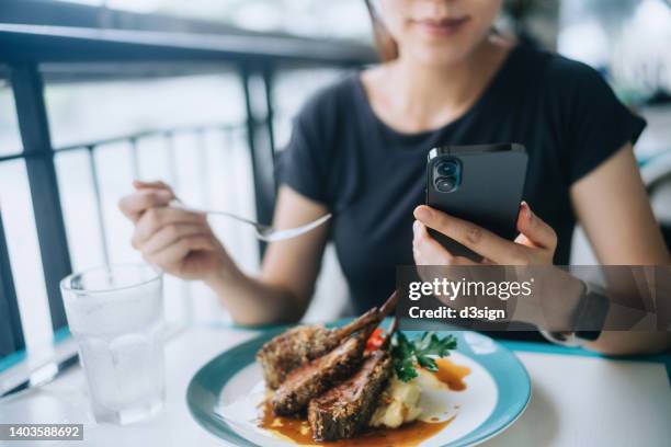 cropped shot of young asian woman sitting by the window, using smartphone while enjoying lunch at fine dining restaurant. people, food, lifestyle and technology concept - steakhouse stock-fotos und bilder