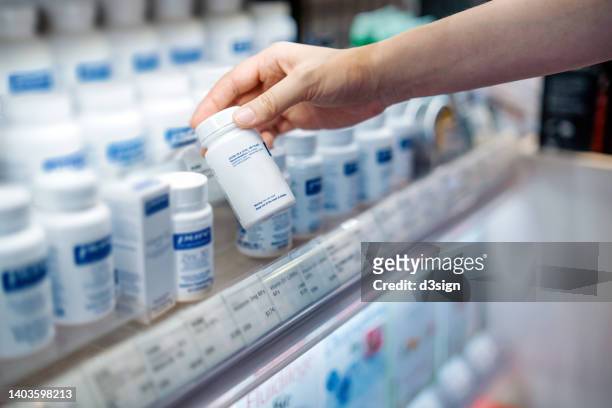 cropped shot of young asian woman's hand taking a bottle of medicine from the shelf at the pharmacy, reading the product information. healthcare, medication and people concept - herbal medicine foto e immagini stock