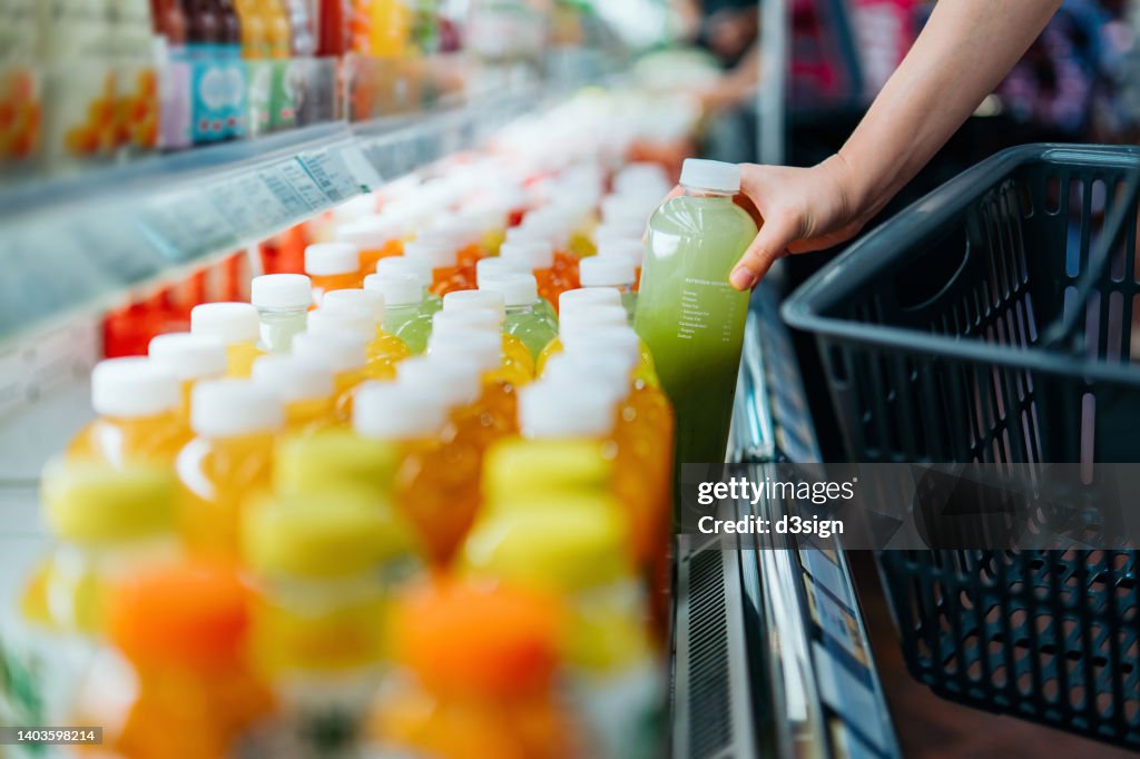 Cropped shot of young Asian woman shopping for fresh fruit juice from refrigerated shelves in supermarket, taking out a bottle of freshly squeezed green juice into a shopping basket. Healthy eating, go green lifestyle