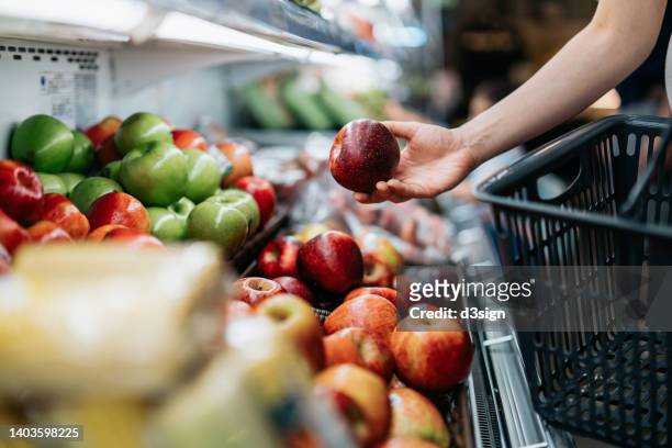 cropped shot of young asian woman choosing fresh organic fruits in supermarket. she is picking a red apple along the produce aisle. routine grocery shopping. healthy living and eating lifestyle - apple stockfoto's en -beelden