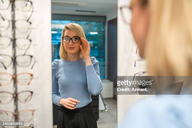 stylish young adult woman trying on glasses and looking into the mirror - optician stockfoto's en -beelden