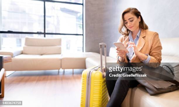 feliz mujer de negocios con equipaje enviando mensajes de texto en su teléfono celular en el vestíbulo de un hotel - huésped fotografías e imágenes de stock