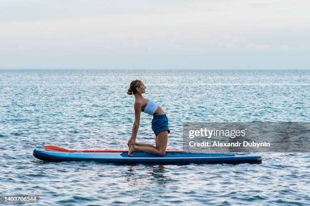 a young woman practices yoga on a sup board against the background of water at the sea in summer - pilates fotografías e imágenes de stock