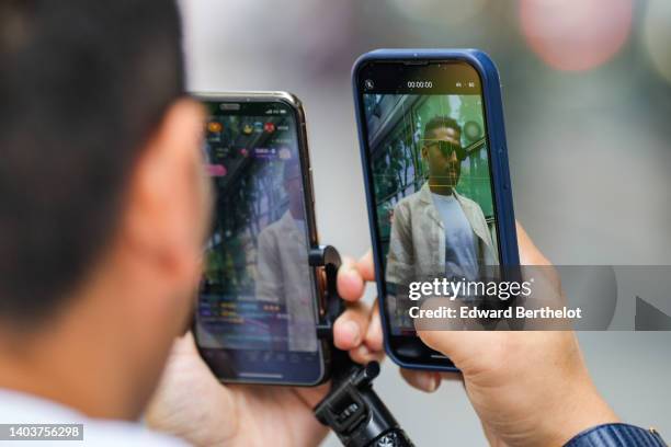 Fan records a live stream video with his smartphone / mobile phone for his social media, and films a guest wearing sunglasses, a white t-shirt, a...