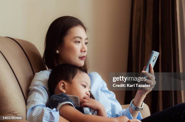 candid portrait of a southeast asian mother using her smart phone in the living room with her son - daily life in manila stock-fotos und bilder