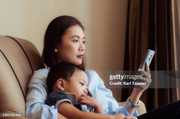 candid portrait of a southeast asian mother using her smart phone in the living room with her son - daily life in manila stock pictures, royalty-free photos & images