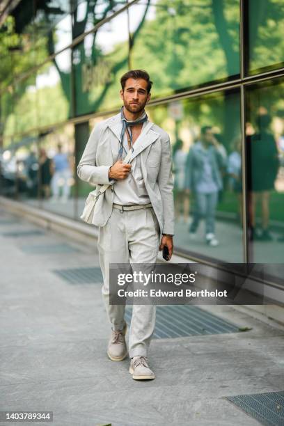 Daniel Toni Jais wears a dark gray and beige print pattern silk small scarf, a pale gray linen blazer jacket, matching pale gray linen suit pants, a...