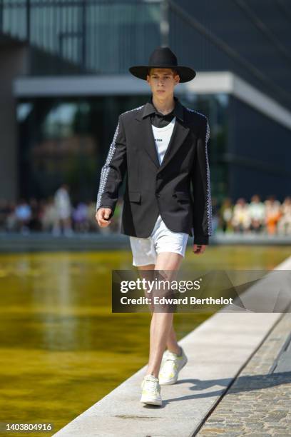 Model wears a black large felt hat, a black shirt from MSGM, a white with black inscriptions print pattern tank-top from MSGM, a black with ethnic...