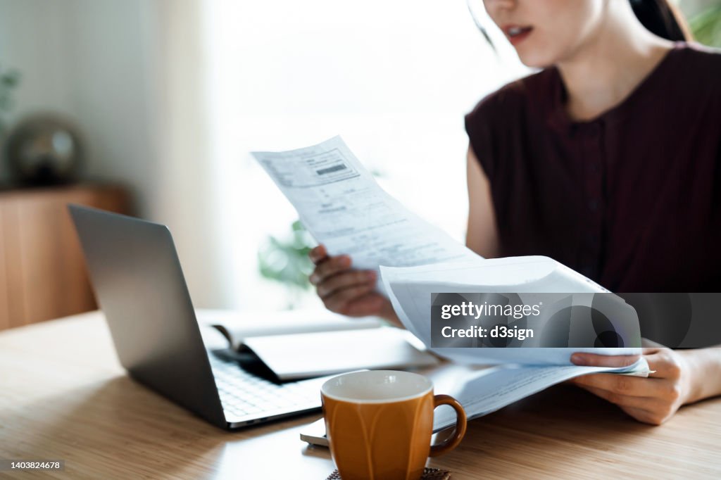 Cropped shot of Asian woman sitting at dining table, handling personal finance with laptop. She is making financial plan and planning budget as she go through her financial bills, tax and expenses at home. Wealth management, banking and finance concept