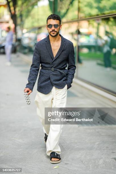 Guest wears sunglasses, a black tweed blazer jacket, a black shiny leather braided belt, white large pants, black suede belted sandals / shoes,...