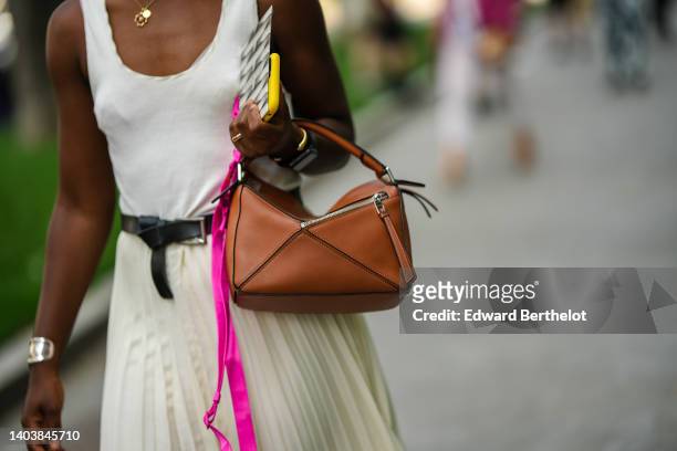 Guest wears a gold chain pendant necklace, a white tank-top, a black shiny leather belt, a camel shiny leather Puzzle handbag from Loewe, a white...