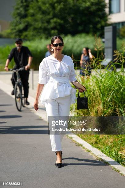 Guest wears black sunglasses, a white ruffled blouse with a large belt, white denim cigarette pants, a black shiny leather handbag, black velvet...