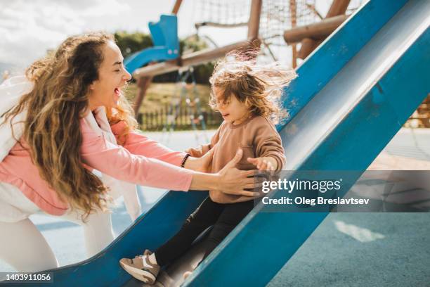 laughing mom catching her happy daughter sliding on kids playground - playground stock photos et images de collection