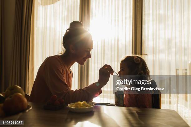 happy mother feeding little baby child with beautiful sunlight in room from window. family harmony - baby spielt mit essen stock-fotos und bilder